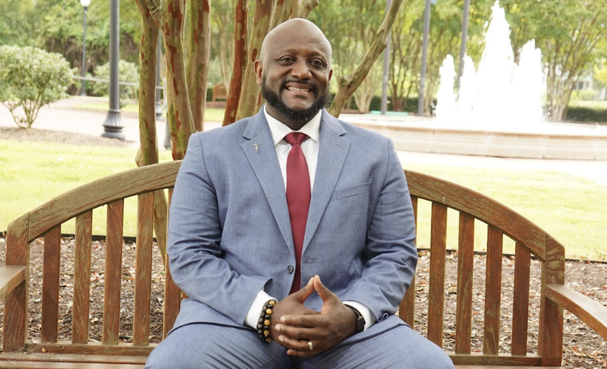 Dr. Curtis Williams seated on a park bench, smiling, with a fountain behind him